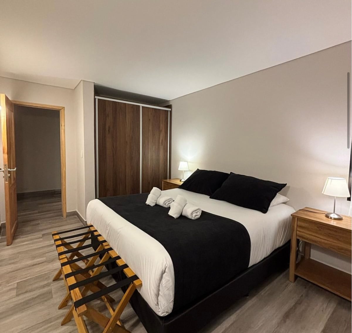 A well-appointed bedroom features a spacious bed with dark bedding and white pillows. Nightstands with lamps are positioned on either side. A wooden wardrobe complements the neutral-colored walls, while a small bench is placed at the foot of the bed. Light-colored flooring adds to the calm ambiance.