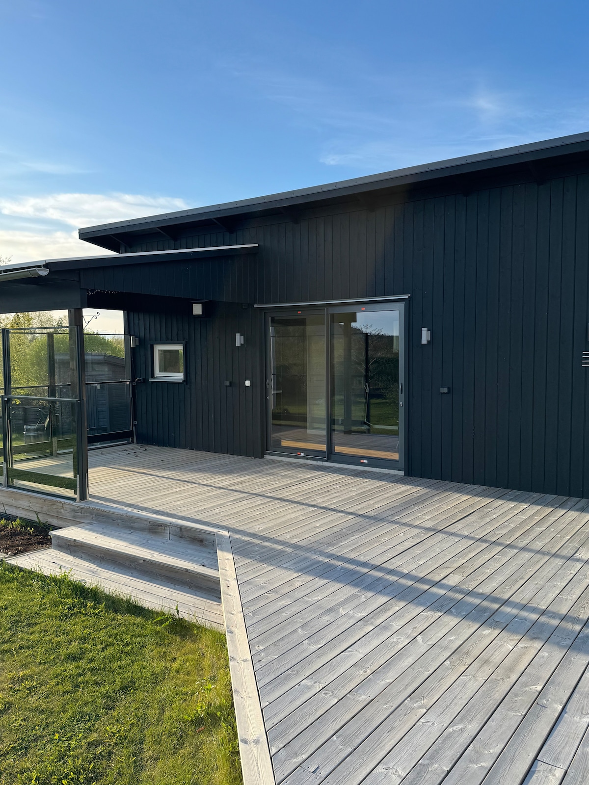 The exterior of the guest house features a modern black wooden facade with large glass doors that provide access to a deck. The surrounding area is adorned with green grass, and the steps leading up to the entrance are made of grey wood.