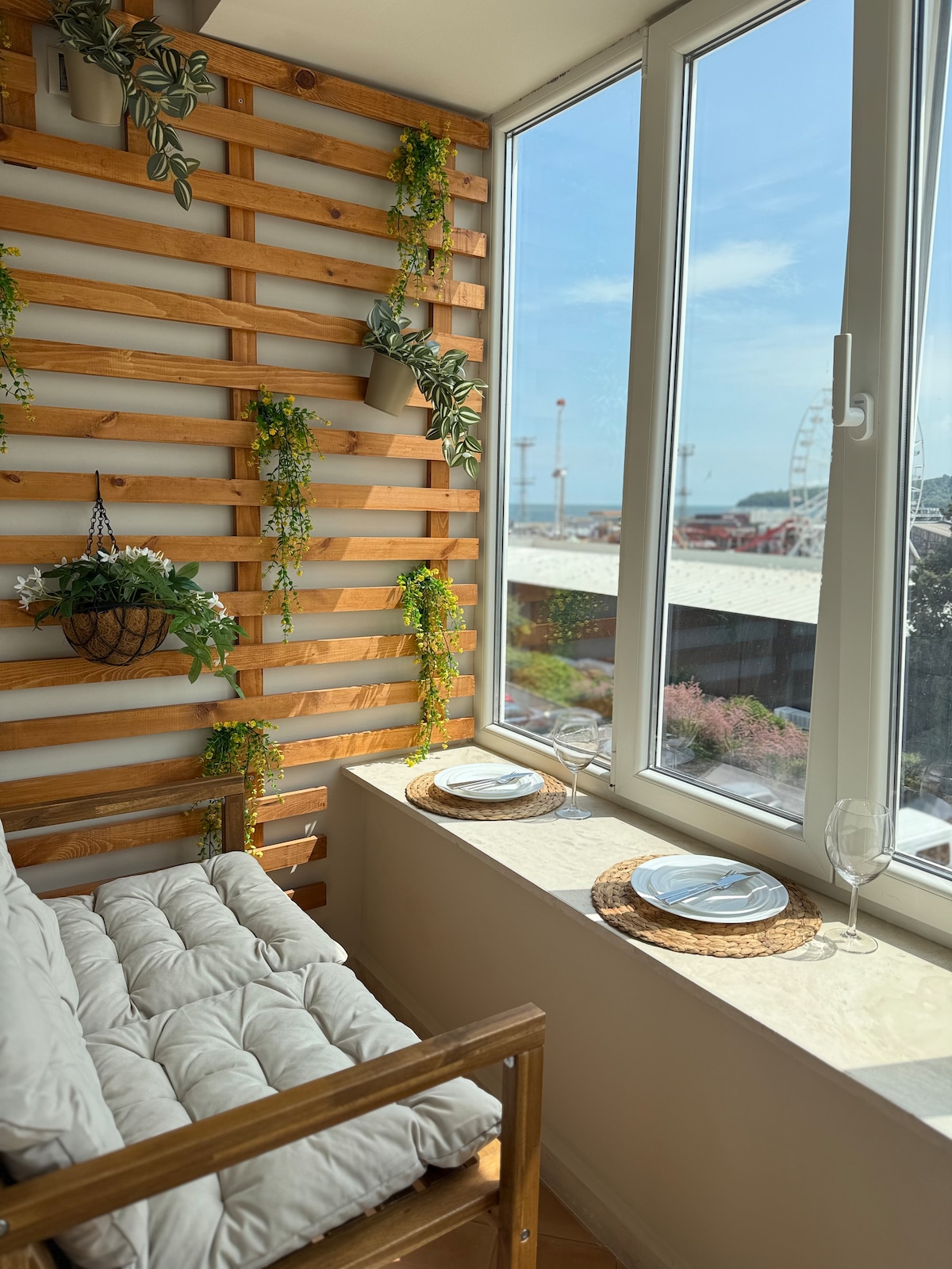 A cozy balcony space features a wooden wall adorned with greenery. Two plates and wine glasses are set on a sunlit window ledge. The view outside includes a glimpse of the sea and nearby attractions, creating a peaceful atmosphere.