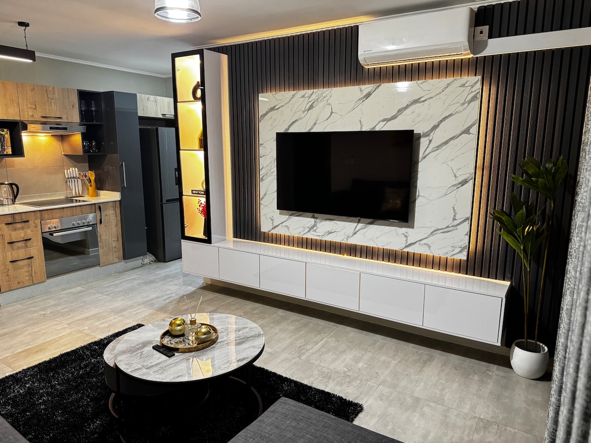 An inviting living area features a modern wall-mounted television against a marble-patterned backdrop, complemented by ambient lighting. A stylish coffee table sits atop a dark area rug, while a potted plant adds a touch of greenery to the space. The open layout connects to the kitchen area.