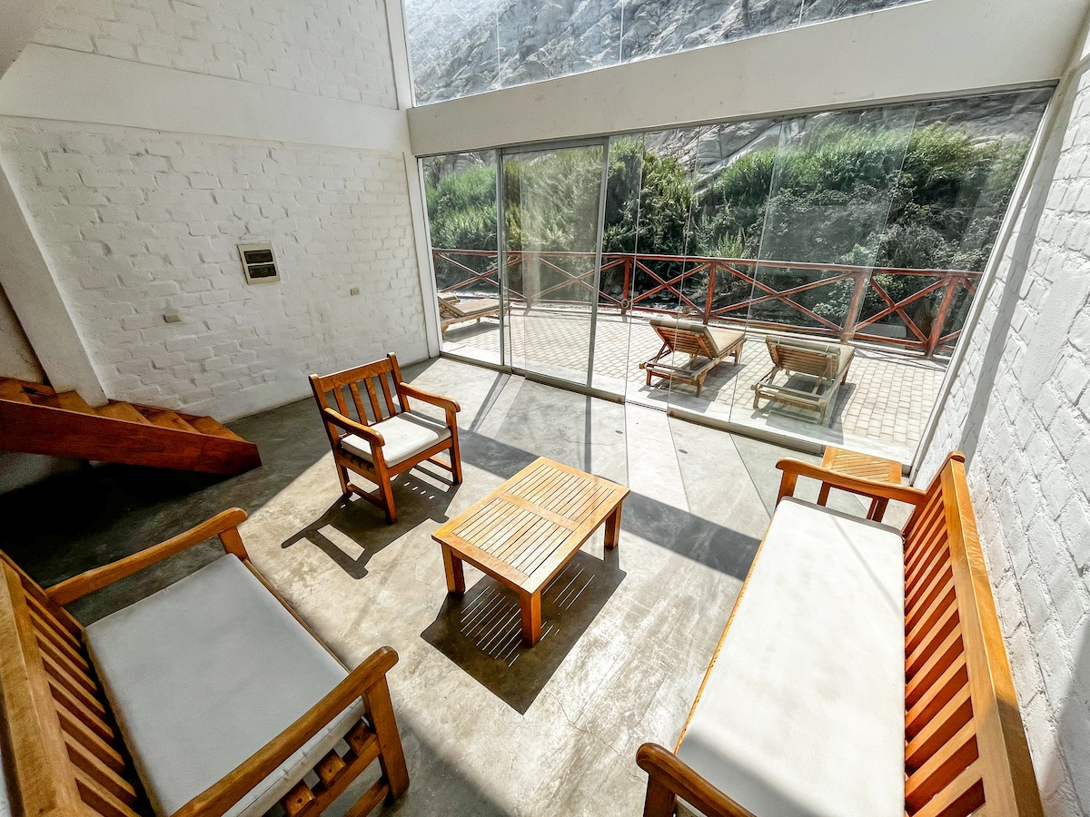 A bright living area features large glass panels that open to a view of the surrounding greenery. Wooden furniture, including two armchairs and a matching table, is arranged on a polished concrete floor, providing a comfortable space for relaxation and connection with nature.