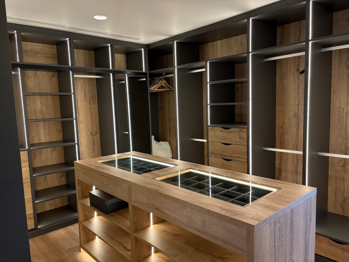 A spacious walk-in closet features numerous shelves and storage options with a sleek design. A central island is highlighted with a glass top and under-shelf lighting, and wooden textures provide a warm aesthetic.