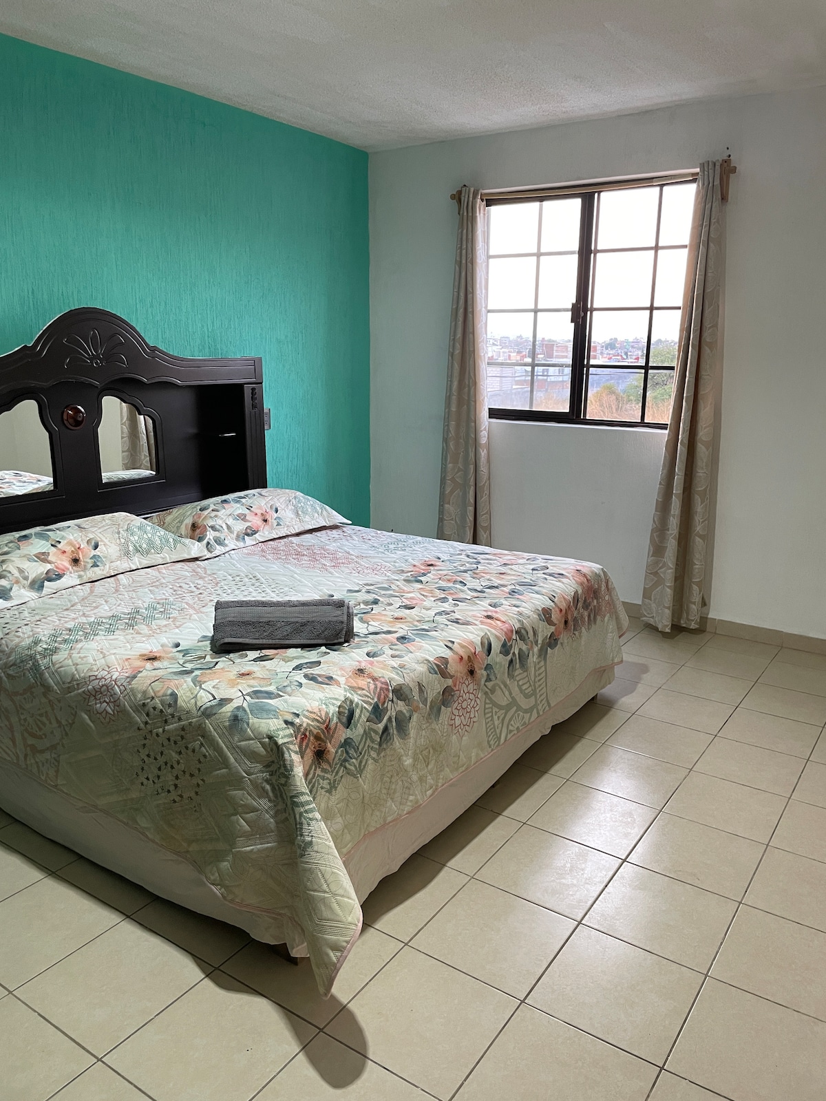 A bright sleeping area features a spacious bed with a floral patterned quilt and a neatly folded towel. A large window allows natural light to brighten the room, complemented by light curtain panels. A smooth tiled floor enhances the minimalist aesthetic.