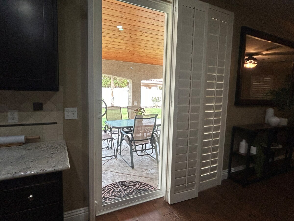 A view from the kitchen leads to a covered outdoor patio, featuring a dining set with multiple chairs. Natural light filters in through the open door, highlighting the inviting space with a wooden ceiling and landscaped yard visible beyond.