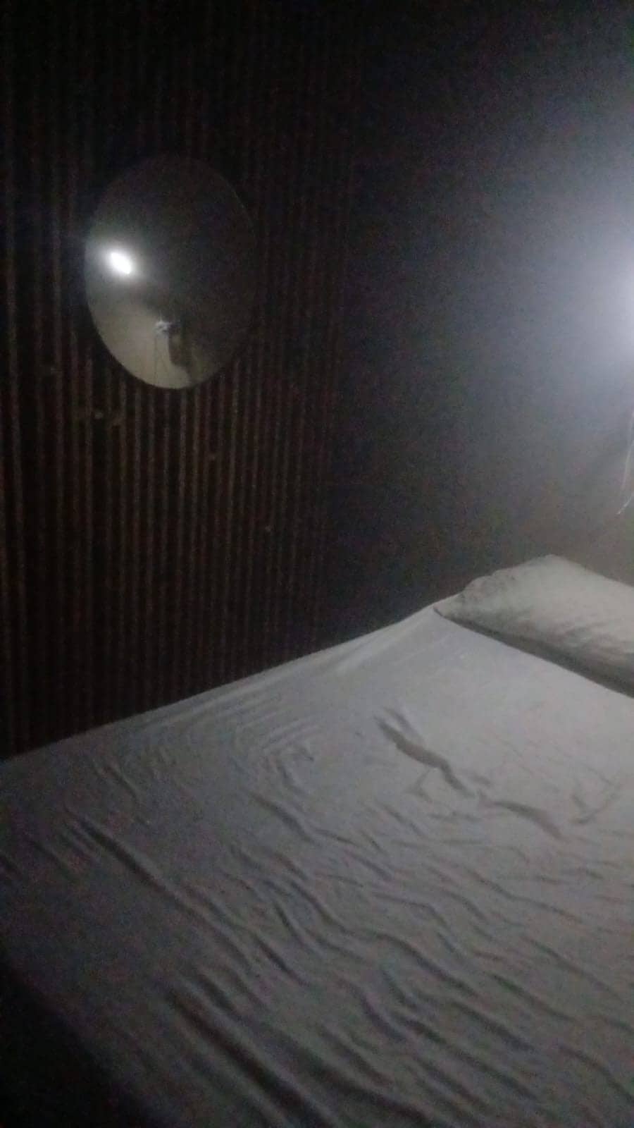 A softly lit bedroom features a spacious bed with a wrinkled white sheet, positioned against a textured wall. A round mirror reflects light from a nearby source, casting a gentle glow over the space.