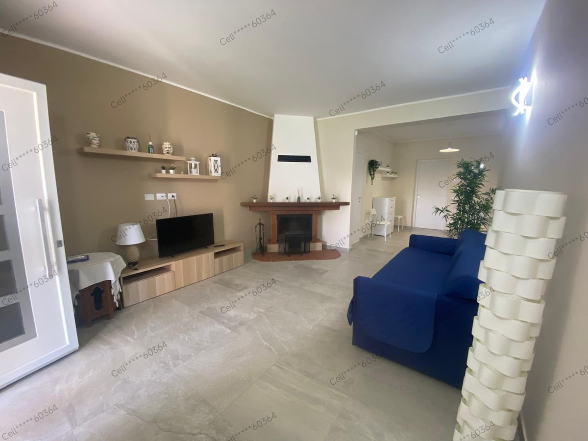 A spacious living room features a comfortable blue sofa facing a wooden fireplace. A television is positioned on a shelf beneath a decorative display. Natural light illuminates the room, highlighting the neutral wall color and modern decor.