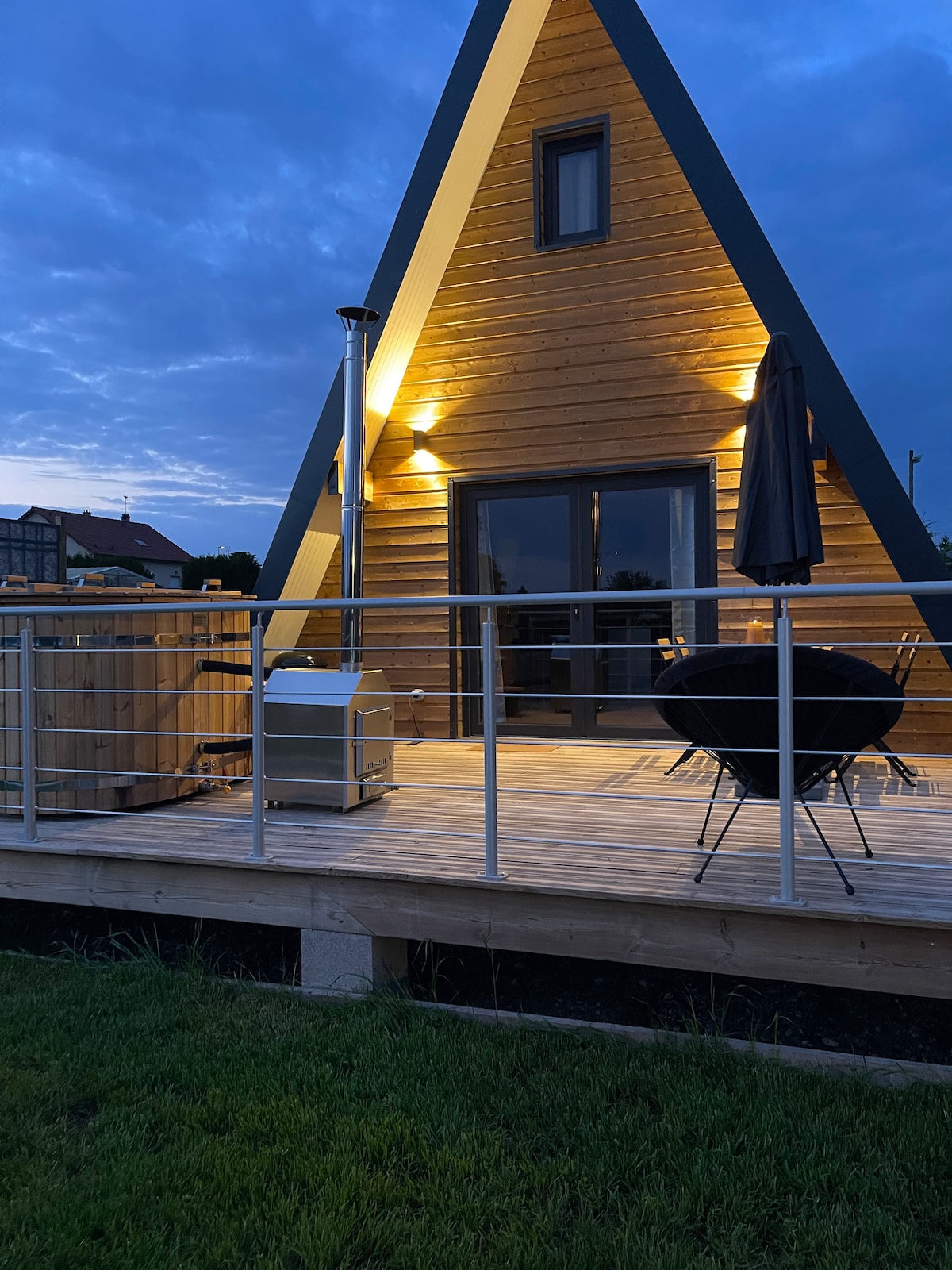 An A-frame lodge is seen under an evening sky, illuminated by warm lights. A spacious deck features a sleek metal outdoor heater and a round black chair. A tall umbrella stands beside the chair, adding to the cozy outdoor experience.