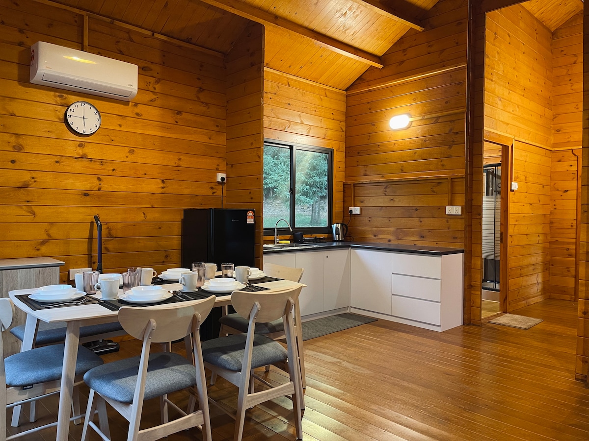 An open-concept kitchen area features wooden walls and a high ceiling, with a dining table set for four. Kitchen appliances include a refrigerator and kettle, while natural light enters through a window. A modern bathroom is visible through an adjacent doorway.