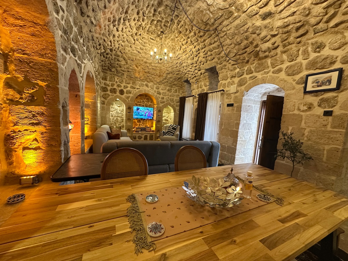 The rustic interior features a high arched stone ceiling and beautifully crafted stone walls. A wooden dining table is positioned in the foreground, complemented by comfortable seating nearby. Soft lighting creates an inviting atmosphere, while a television is visible in the background, framed by decorative wall arches.