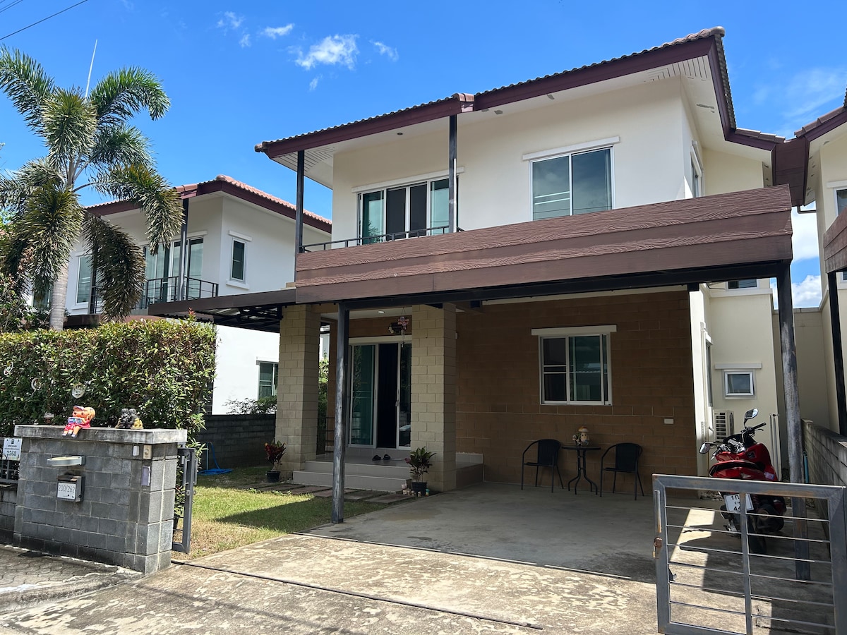 The exterior of the two-story house features a welcoming porch with two chairs, surrounded by well-maintained landscaping. Large windows provide ample natural light, while palm trees add greenery to the scene. A motorcycle is parked on the paved area near the entrance.