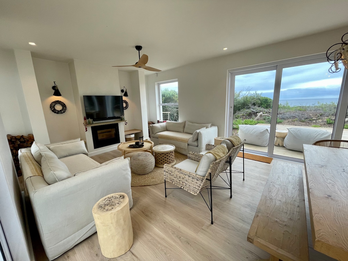 A comfortable living area is presented, featuring soft seating arrangements with neutral tones. A flat-screen television is mounted on the wall, and natural light streams through large glass doors that offer views of the surrounding landscape. A rustic dining table and elegant light fixtures add charm.