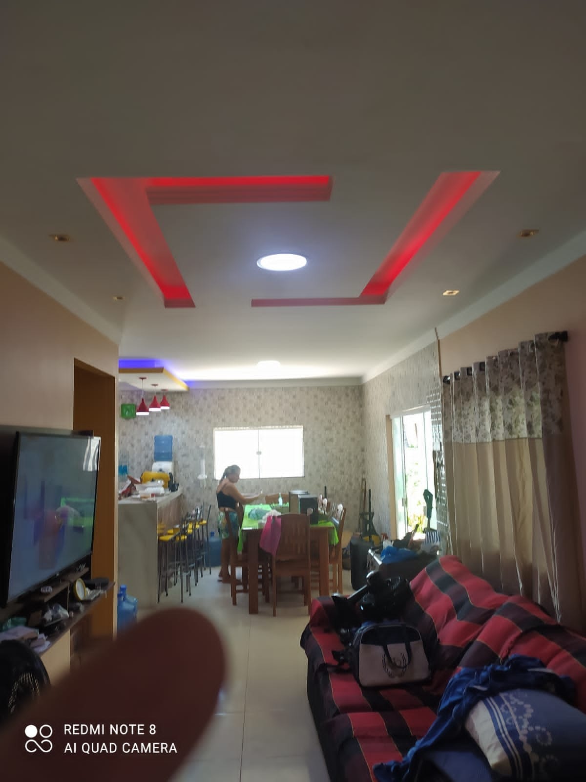 An open living area showcases a modern design with recessed lighting highlighting the ceiling's geometric pattern. A comfortable seating arrangement includes a dark sofa with red and white stripes. Dining tables and chairs are visible in the background, along with natural light streaming through large windows.