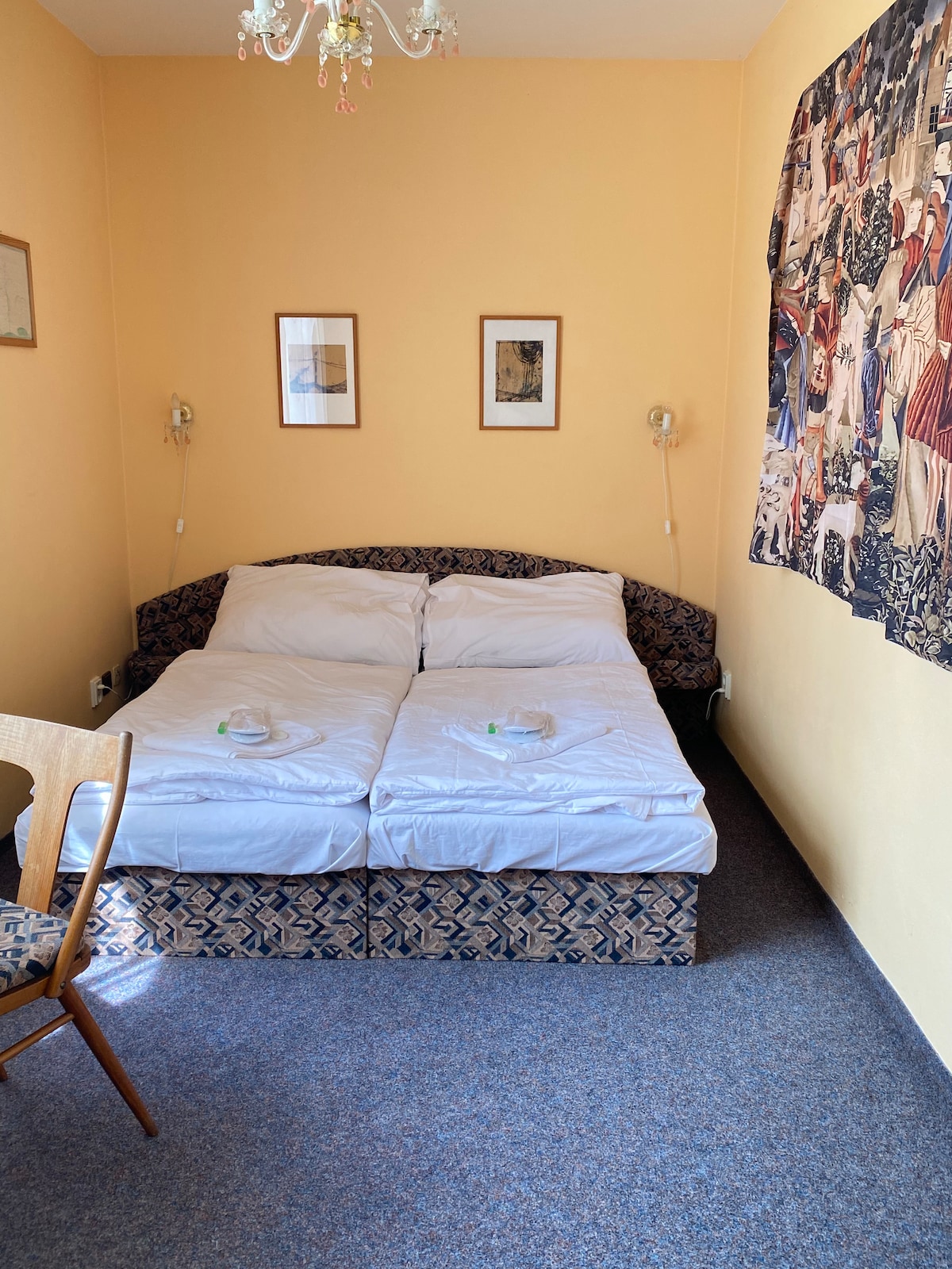 A cozy sleeping space features a twin bed with separate mattresses covered by white linens. Two framed artworks hang above the bed, while a colorful tapestry adorns one wall. A simple wooden chair and table are positioned nearby, providing functional decor.