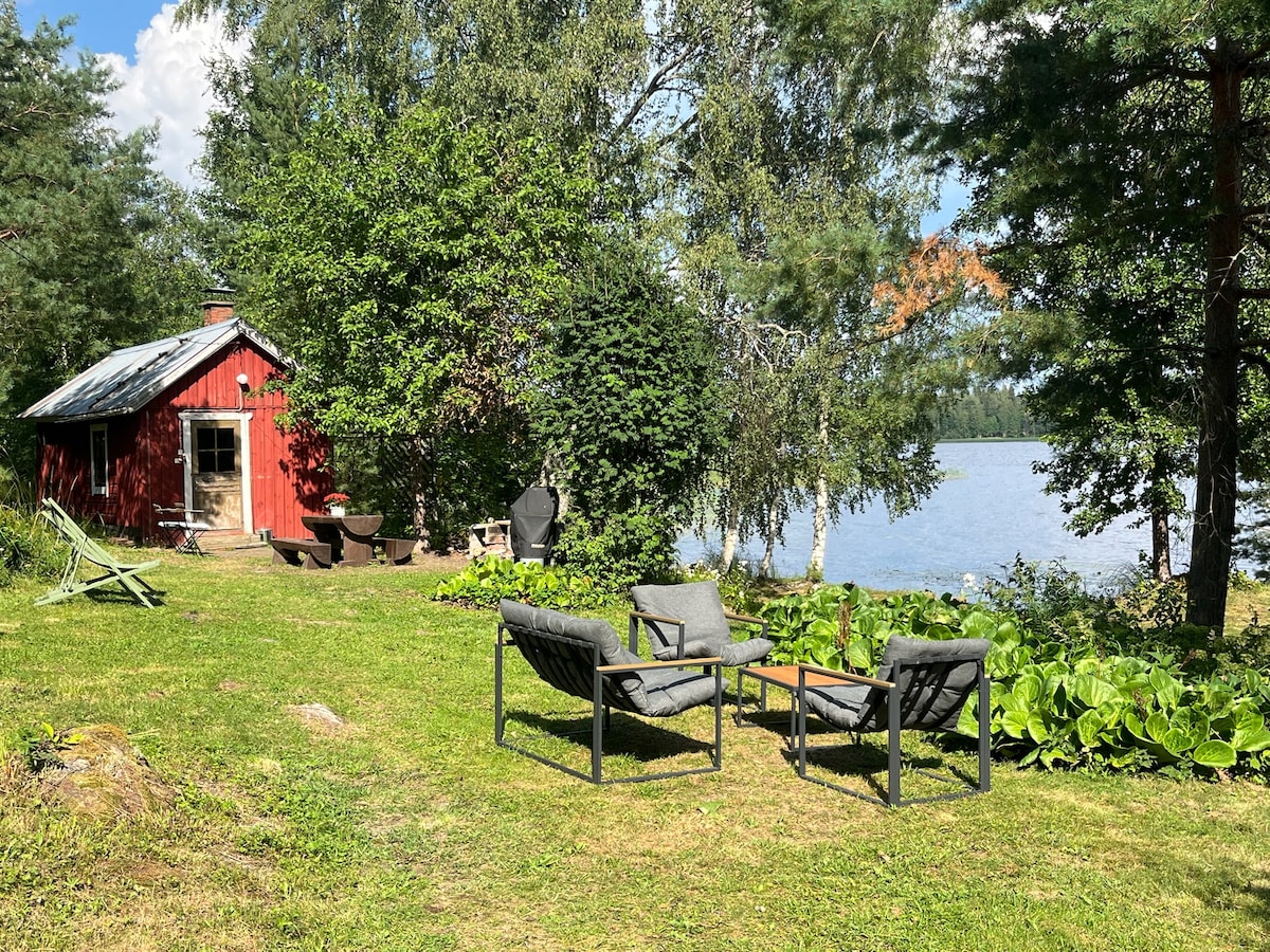 A serene outdoor space features a grassy area with two comfortable chairs and a small table. A cozy red cabin is visible in the background, surrounded by trees and plants. The calming waters of Päijänne are seen nearby, creating a tranquil atmosphere.