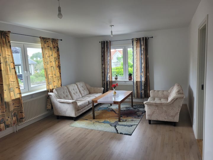 غرفة معيشة، نافذتان كبيرتان مع ستائر سميكة، أريكة بخمسة مقاعد، طاولة وسجادة، مصباح