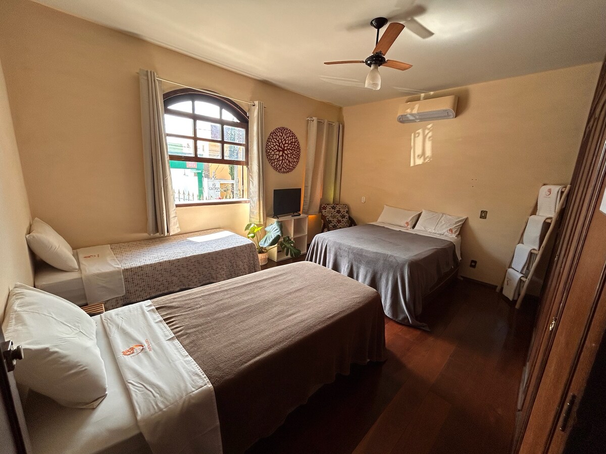 A spacious guest room is presented with three beds, including two twin beds and one larger bed. Natural light filters through an arched window, illuminating the soft-colored walls. A small television and decorative items are visible, and a ceiling fan is installed for comfort.