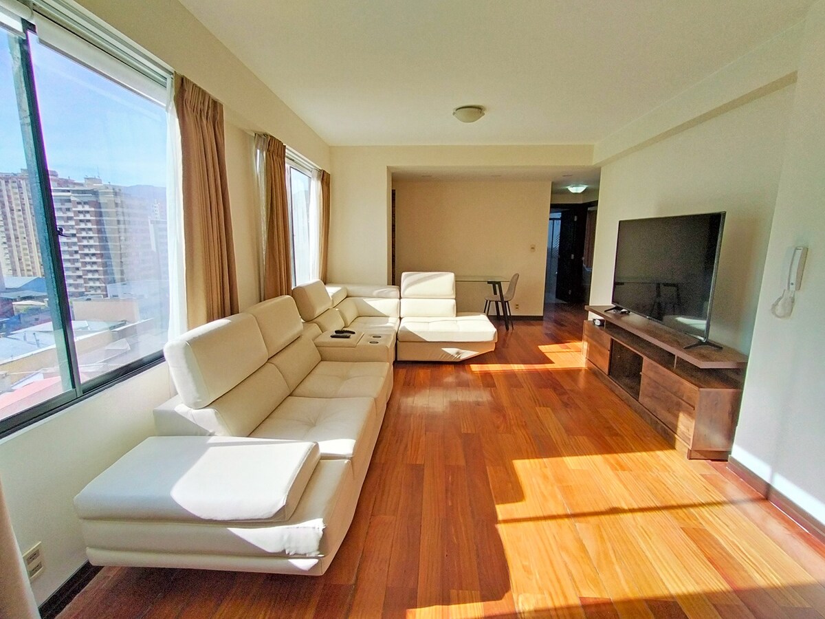 A spacious living area is illuminated by natural light from large windows, showcasing a light-colored sectional sofa. A modern TV is positioned on a wooden media console, and a small chair can be seen in the background among the warm wooden flooring.