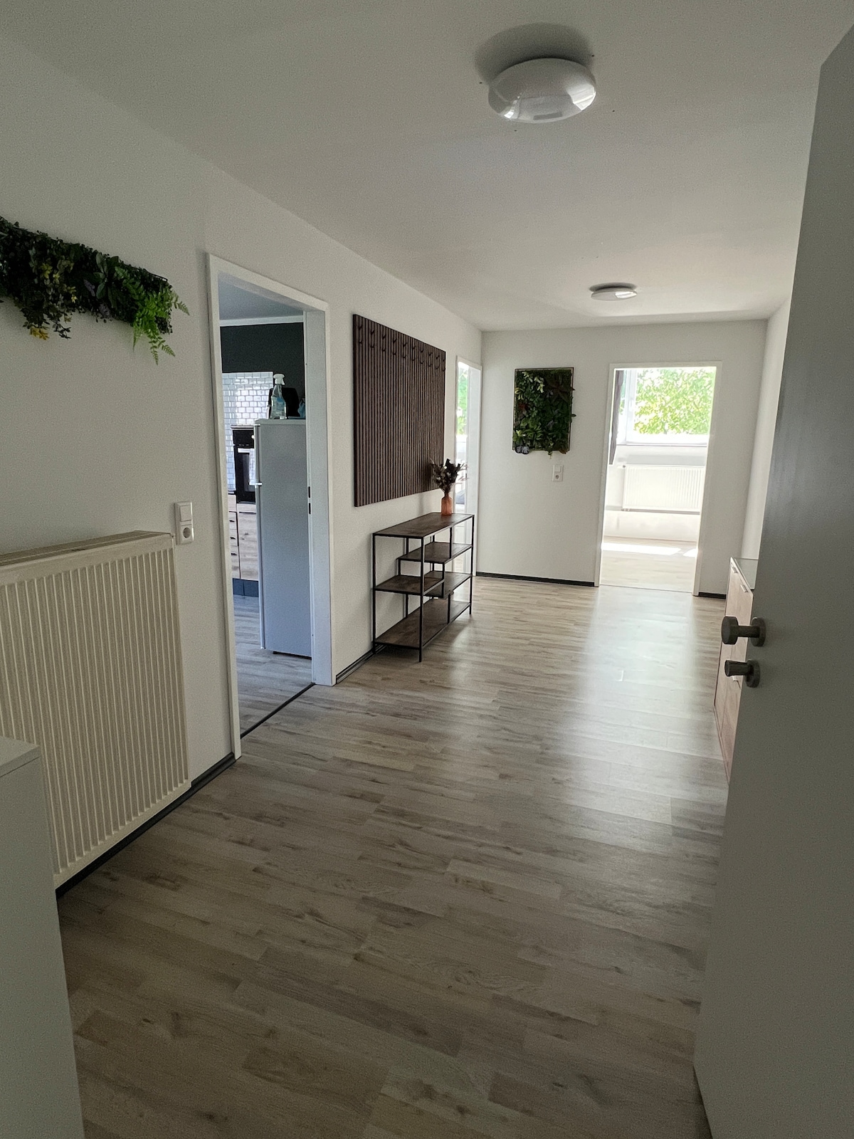 A bright hallway is depicted with light-colored walls and a wooden floor. Decorative greenery adorns the walls, complementing the minimalist decor. A small shelf is positioned against one wall, while doorways lead to other rooms, enhancing the open and airy feel of the space.