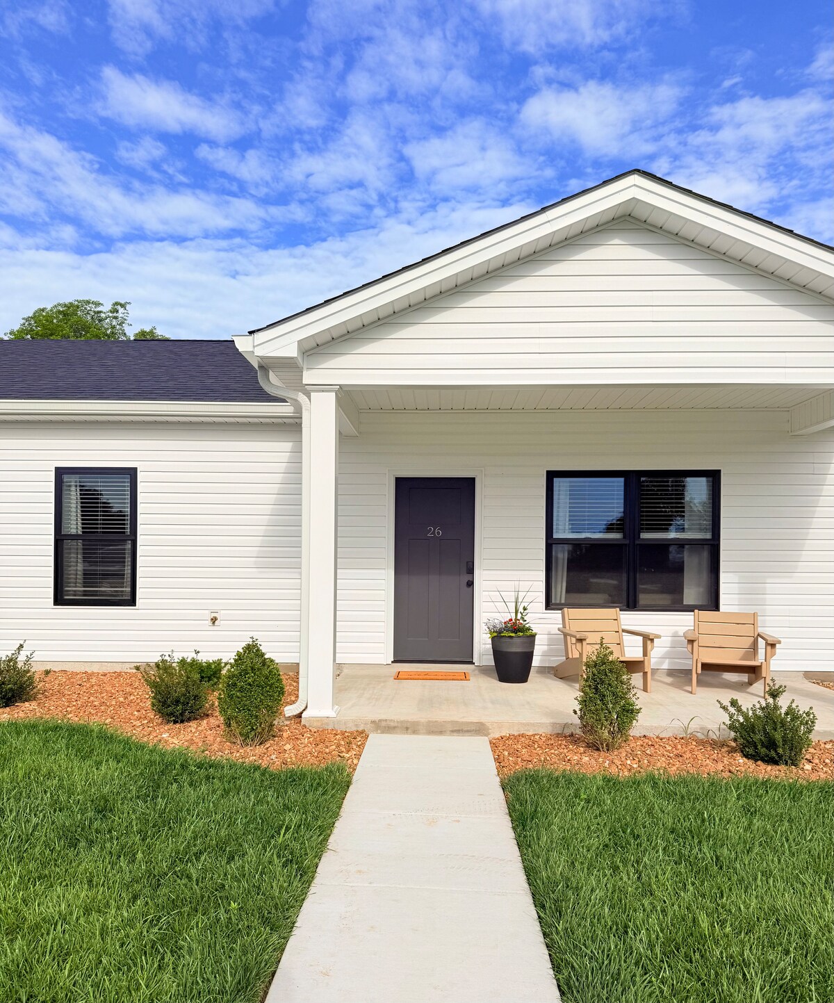 The entrance features a covered porch with two wooden chairs positioned beside a potted plant. A concrete walkway leads to the front door, flanked by fresh green grass and well-maintained landscaping, complemented by light gravel details.
