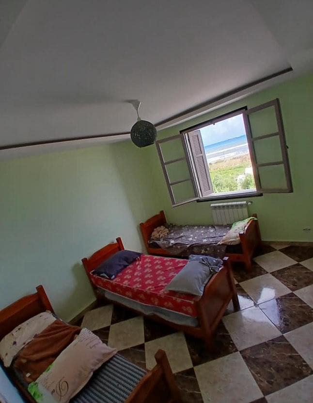 A serene bedroom is presented with three wooden beds, each featuring different bedding. Natural light enters through a window, offering a view of the outside landscape. Light green walls and tiled flooring create a calming environment.