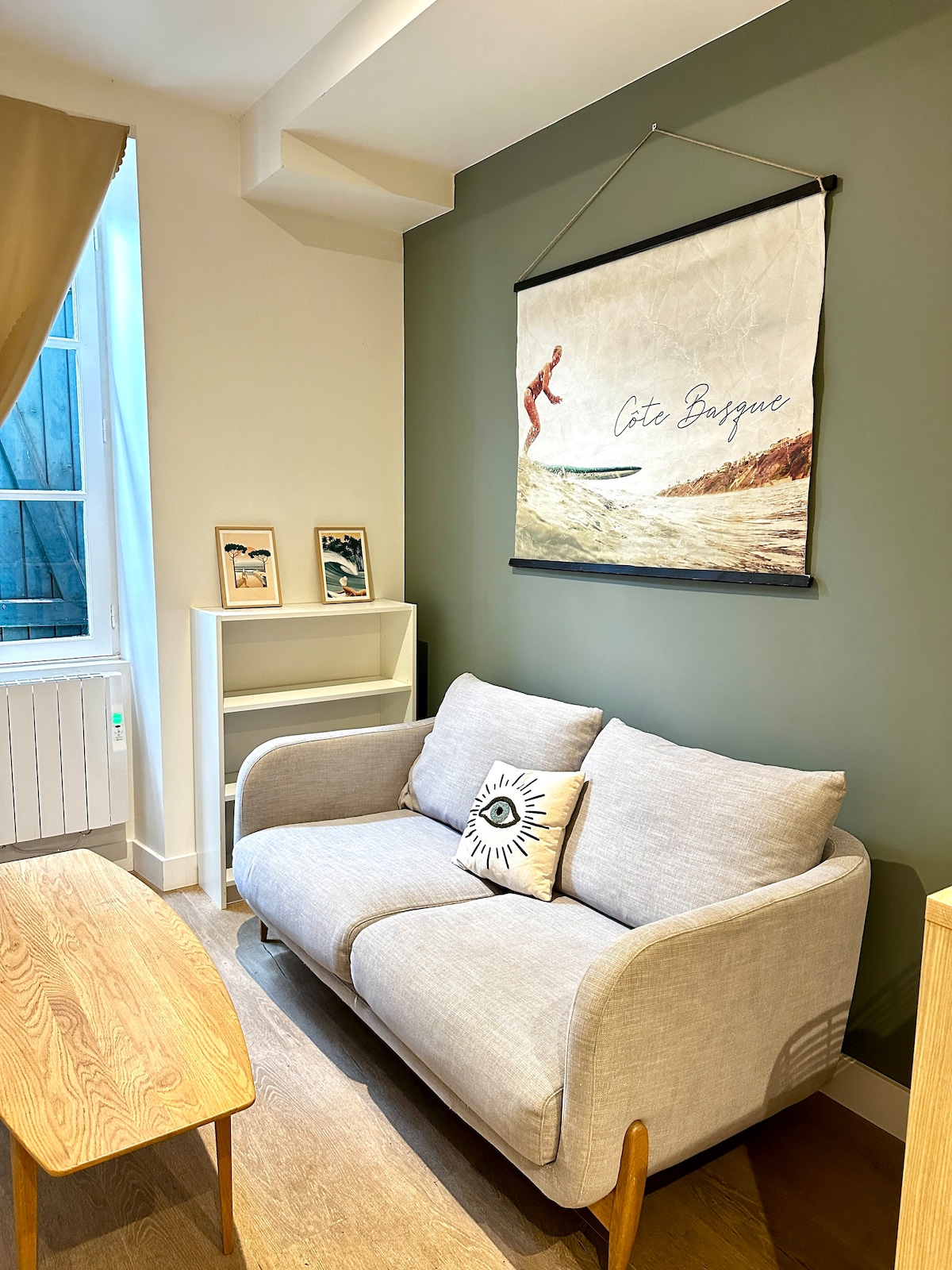 A light-filled living area contains a comfortable grey sofa with a decorative pillow featuring an eye motif. A wooden table sits in front of the sofa. A wall-mounted artwork depicts a surfer and the words 'Côte Basque,' accented by two framed pictures on a shelf nearby.