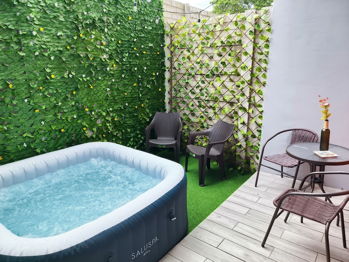 An outdoor space features a cushioned inflatable hot tub surrounded by a lush green wall adorned with artificial foliage. Two wicker chairs and a small round table are positioned on a smooth, light-colored deck. A decorative flower arrangement adds a touch of color.