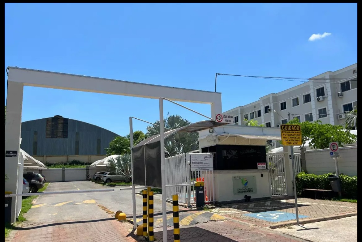 The entrance to the property is depicted, featuring a security gate and a small guard booth. Clear weather enhances visibility of the surrounding buildings, with a large structure visible in the background. Yellow caution bollards are positioned along the drive, adding to the secure atmosphere.