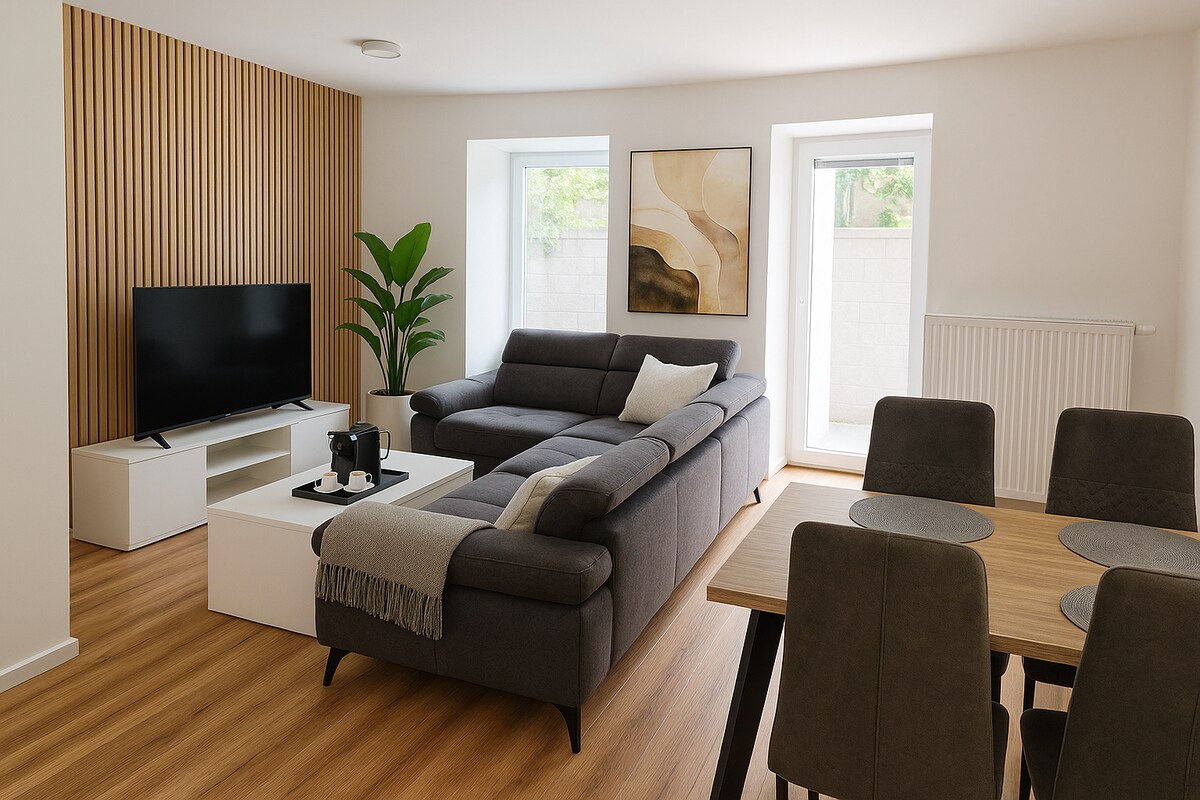 A lounge area features a modern sectional sofa and a coffee table, complemented by a television on a white stand. A dining table with grey chairs is visible. Large windows allow natural light to fill the space, while a decorative plant adds a touch of greenery.