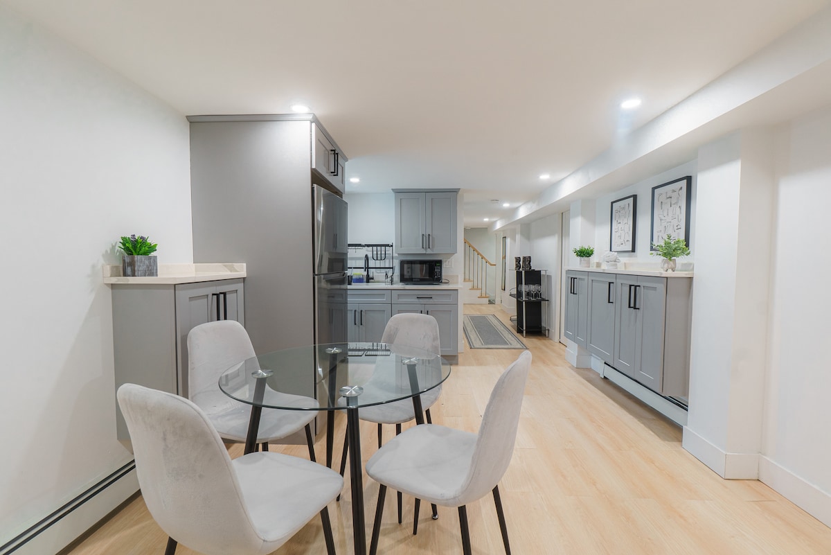 A modern kitchen area is highlighted by sleek gray cabinetry and a dining table with four upholstered chairs. A glass-top table is positioned near a well-equipped kitchen featuring stainless steel appliances, offering a clean and functional space. Soft lighting enhances the airy atmosphere.