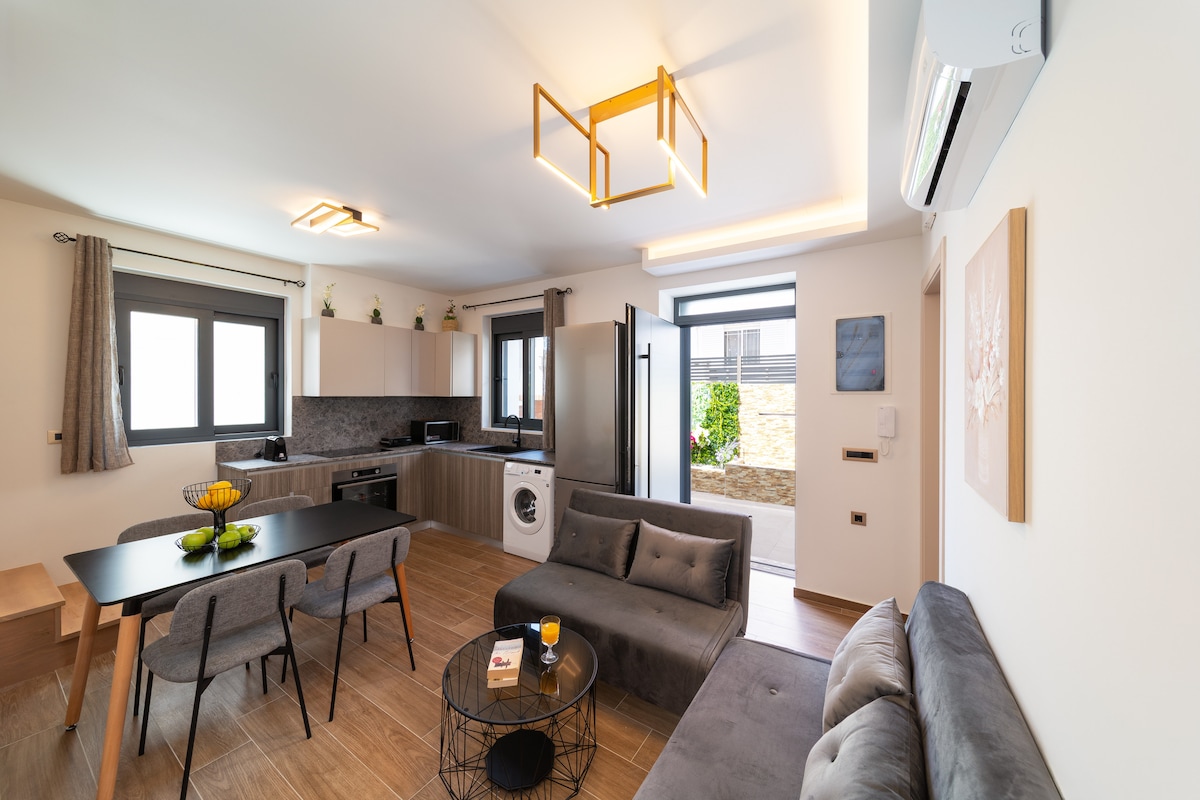 A modern open-plan living space is shown, featuring a comfortable grey sofa and a black dining table surrounded by four chairs. Natural light fills the room through large windows, highlighting the contemporary kitchen with sleek cabinets and a washing machine.