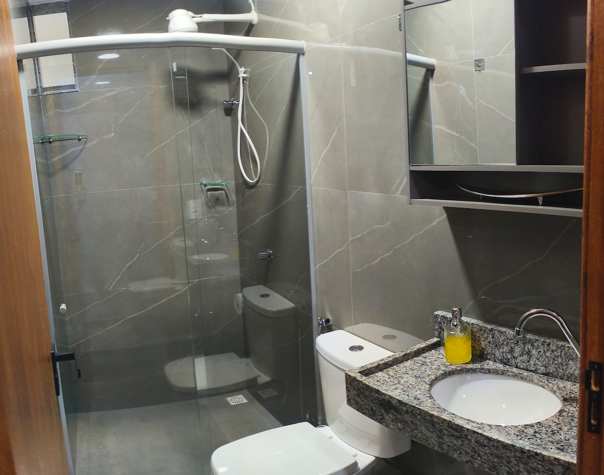 A modern bathroom is featured with sleek grey tiles and a walk-in shower enclosed by glass. A rectangular sink sits atop a granite countertop, complemented by a small mirror above. Essential bathroom amenities are organized neatly, contributing to a clean and functional space.