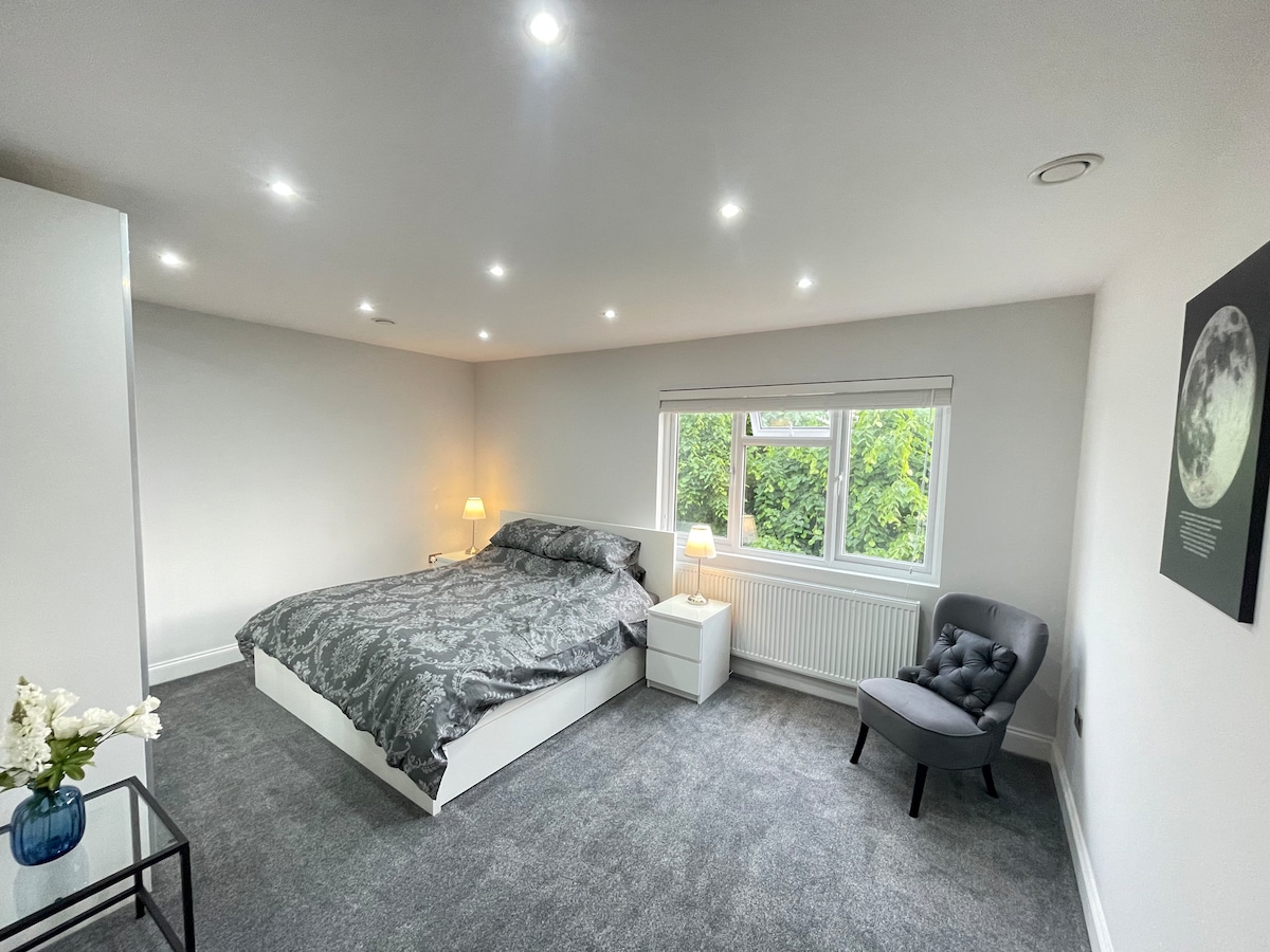 A spacious bedroom is furnished with a large bed covered in a patterned duvet. A bedside lamp provides soft lighting beside a white nightstand. A comfortable armchair is situated in one corner, and a window offers views of green foliage outside.