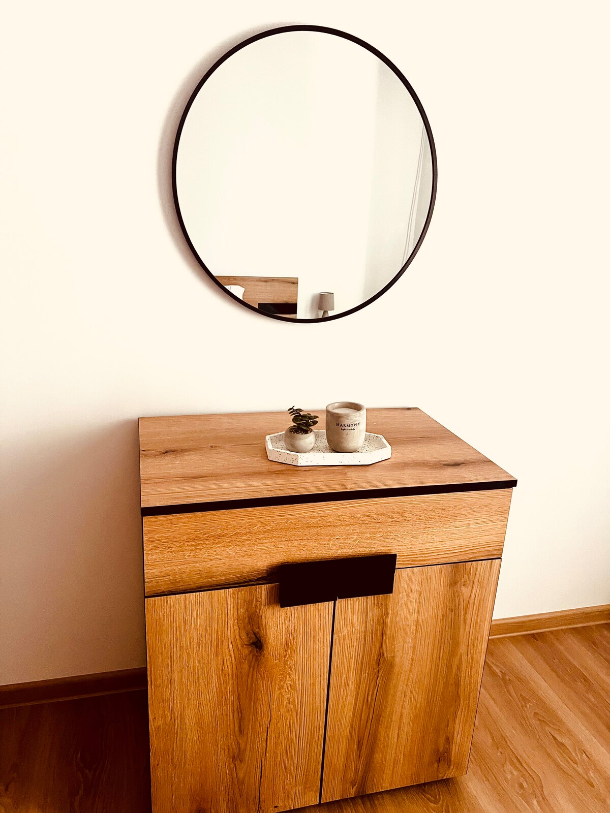 A wooden cabinet is positioned against a light wall, featuring a smooth drawer and a circular black mirror mounted above. A small decorative tray with a candle and a plant sits atop the cabinet, blending functionality with minimalistic design.