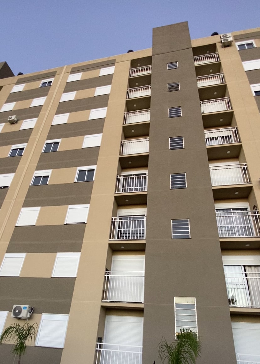 A multi-story residential building is depicted, featuring a modern facade with alternating shades of beige and brown. Balconies with railings are visible, along with multiple windows displaying light-colored shutters. Air conditioning units are mounted prominently on the exterior.