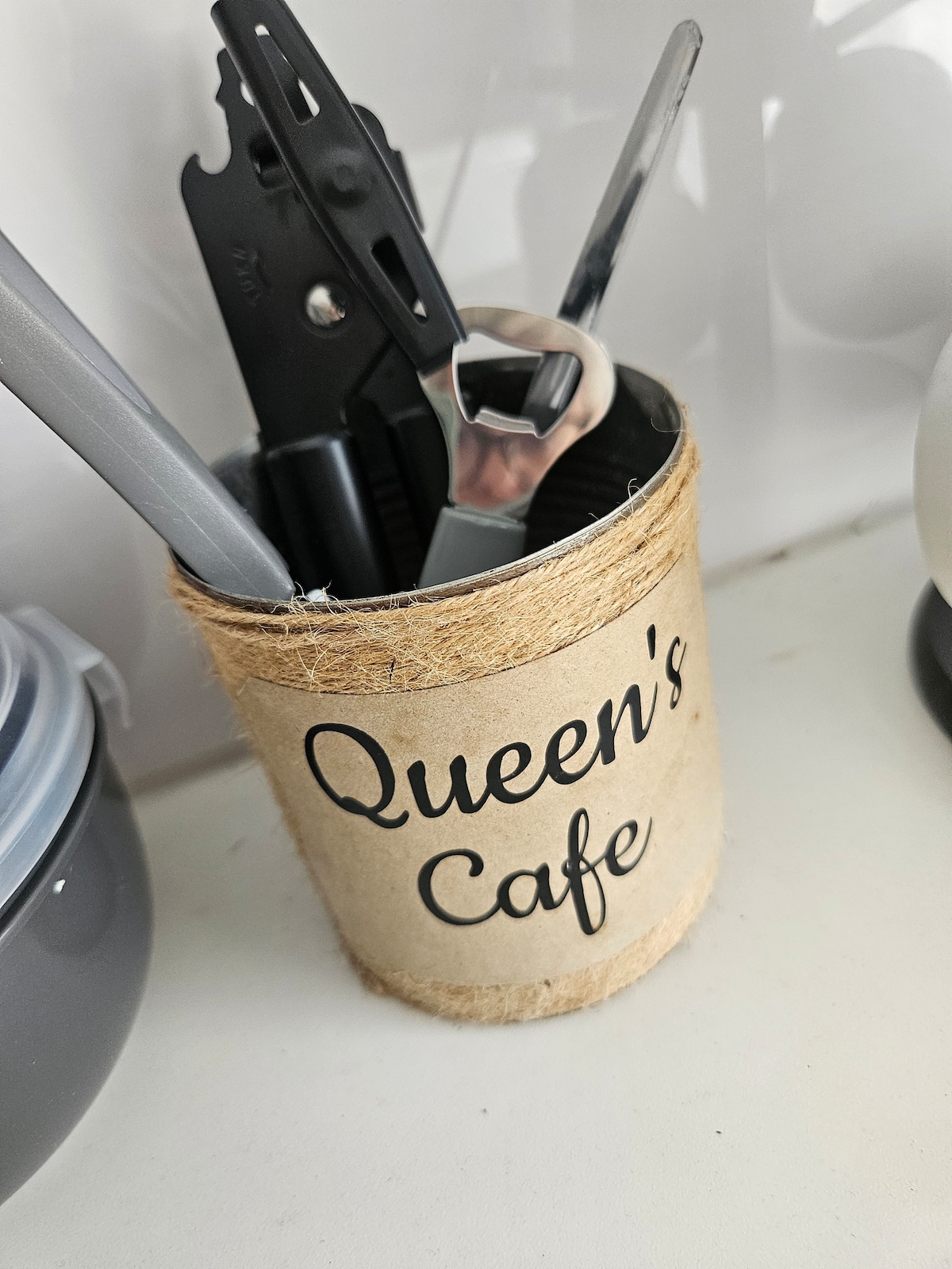 A rustic container labeled 'Queen's Cafe' holds various kitchen utensils, such as a pair of scissors, a can opener, and a couple of spoons. The container is wrapped in jute twine, offering a casual touch to the kitchen decor.