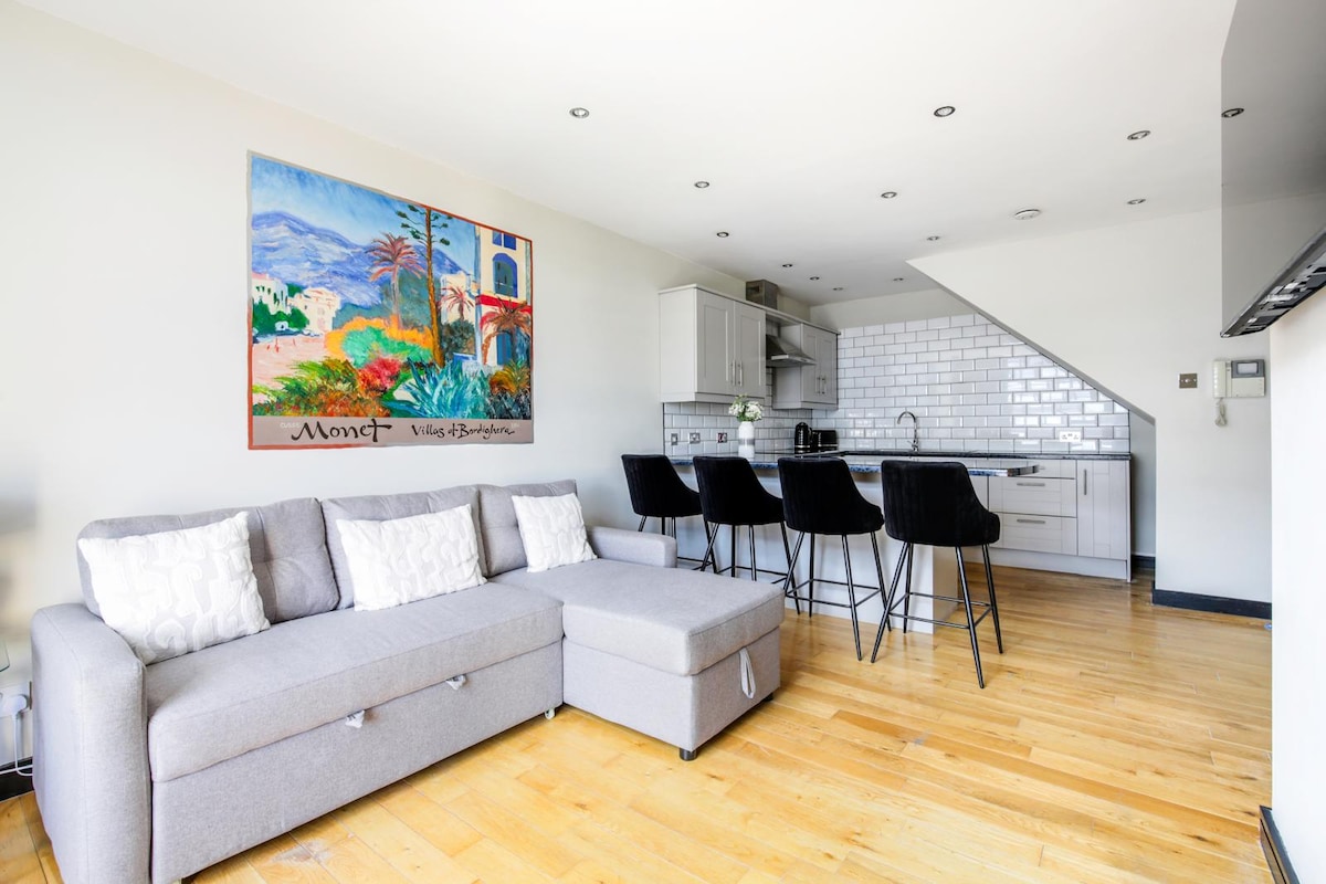 An open-plan living space features a grey sofa with decorative pillows, positioned near a vibrant artwork on the wall. A kitchen area is equipped with modern appliances, including a stovetop and refrigerator. Four black bar stools are arranged around the kitchen island.