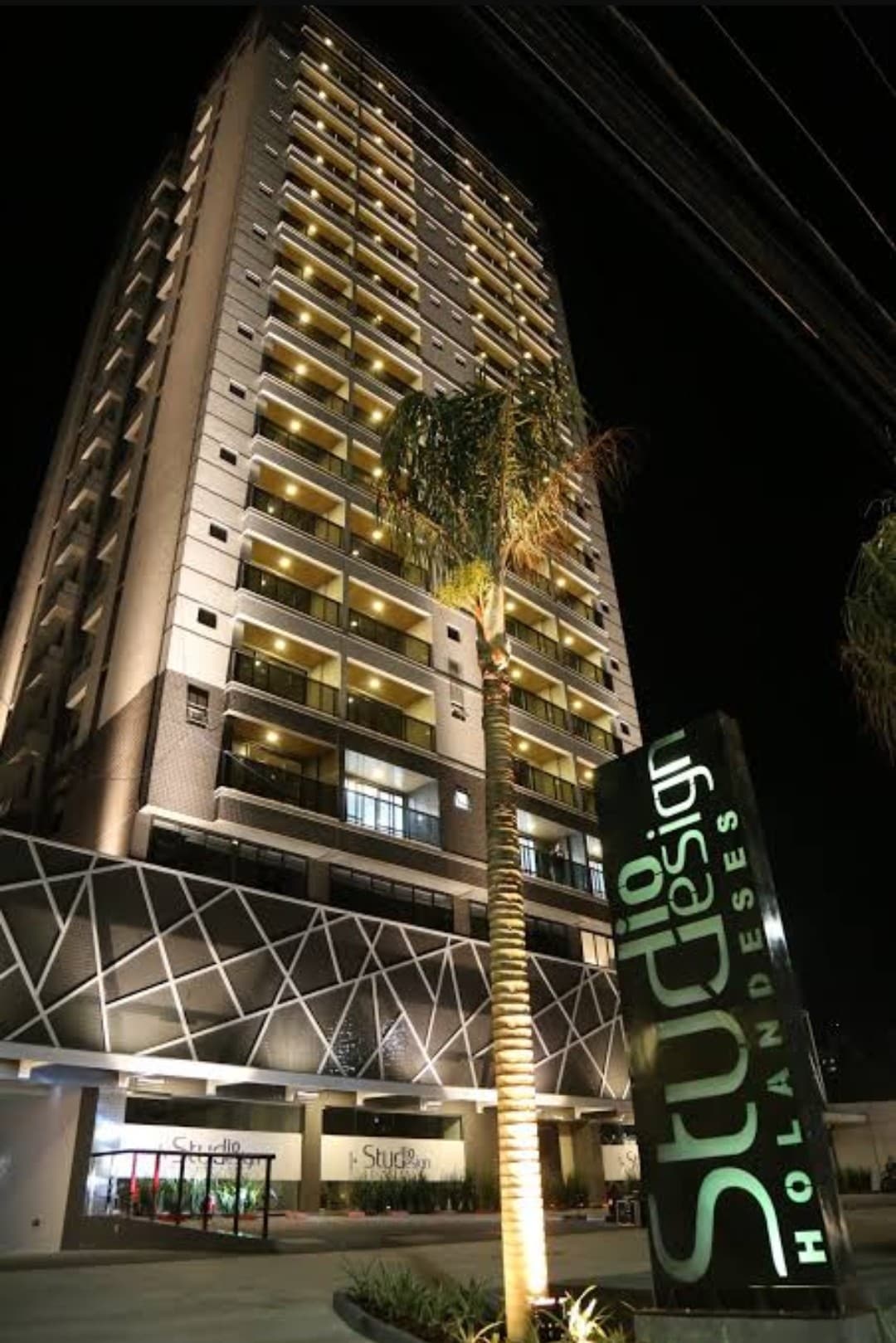 The exterior of the condominium is showcased at night, featuring strong architectural lines illuminated by warm lighting. The building's name, 'Studio Design,' is prominently displayed at the entrance, accompanied by palm trees that add to the modern aesthetic.