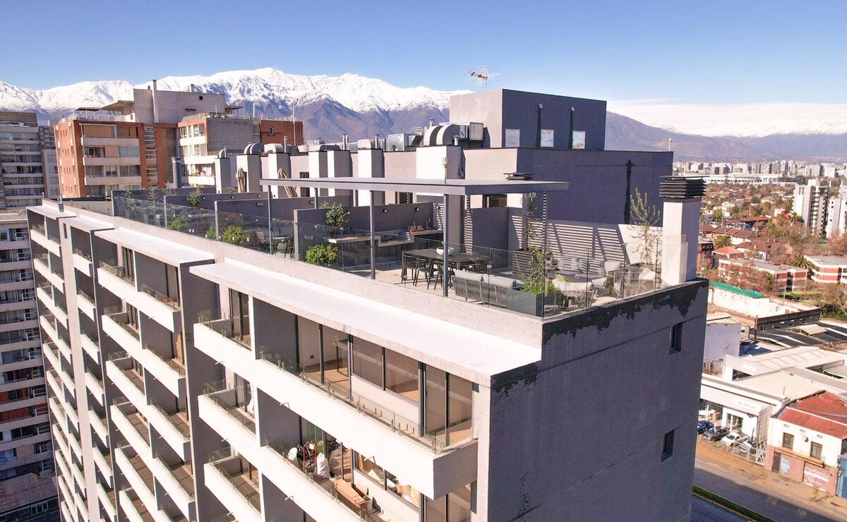 A modern building rooftop is showcased, featuring spacious terraces and plant life. The surrounding views include expansive city landscapes and distant snow-capped mountains, under a clear blue sky. Outdoor seating areas are visible, providing a welcoming space for relaxation.