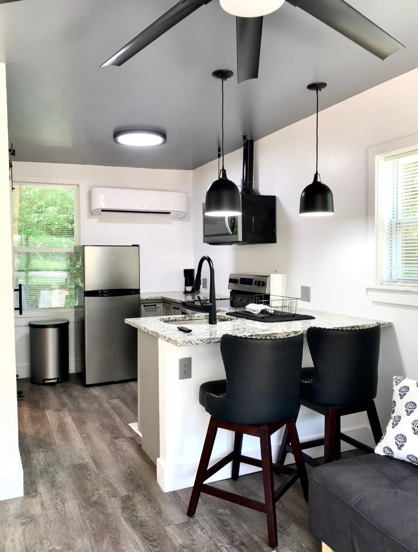 The kitchen area features modern appliances, including a stainless-steel refrigerator and oven, along with sleek black pendant lighting. A compact bar with two dark stools is positioned near a window, creating a bright and functional space. Light hardwood flooring enhances the contemporary design.
