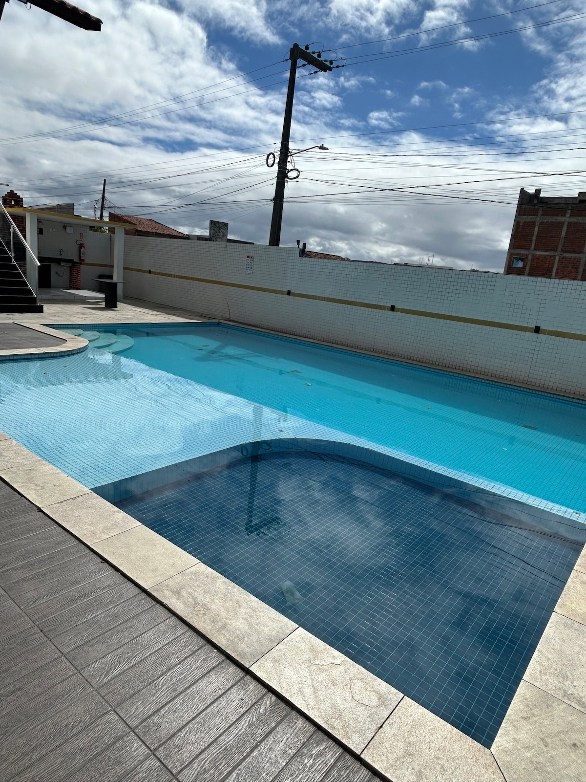 A spacious swimming pool is featured, with clear blue water reflecting the sky. Surrounding the pool is a light-colored tiled area, providing ample space for relaxation. The setting includes a railing, and the background presents a simple wall, contributing to a peaceful environment.