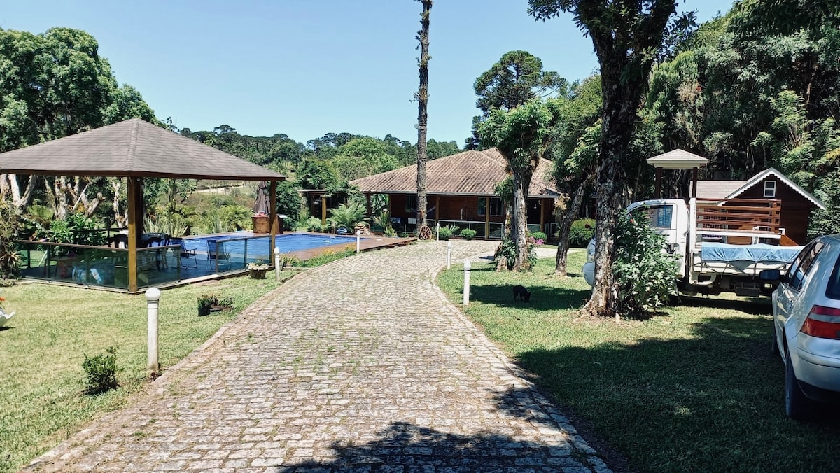 A stone pathway leads through a green lawn towards a wooden structure and a covered outdoor area. A serene body of water is visible on one side, and a few parked vehicles are placed nearby amidst lush trees and open space.