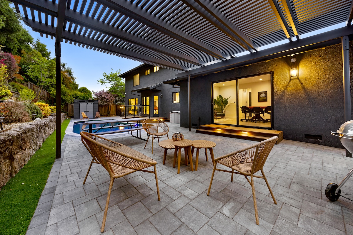 An outdoor seating area features two woven chairs and a round table, set under a slatted pergola. A heated pool is visible to the left, with the modern home’s large glass doors providing a view of the indoor dining space in the background.