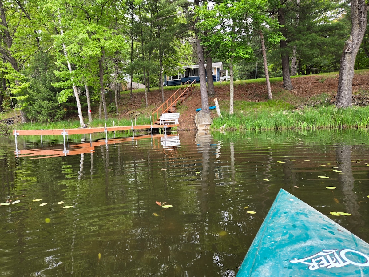 A peaceful lakeside view showcases a private dock leading to a cozy cabin surrounded by lush greenery. The calm waters reflect the surrounding trees, while a kayak's pointed bow is visible in the foreground, inviting exploration of the tranquil lake.