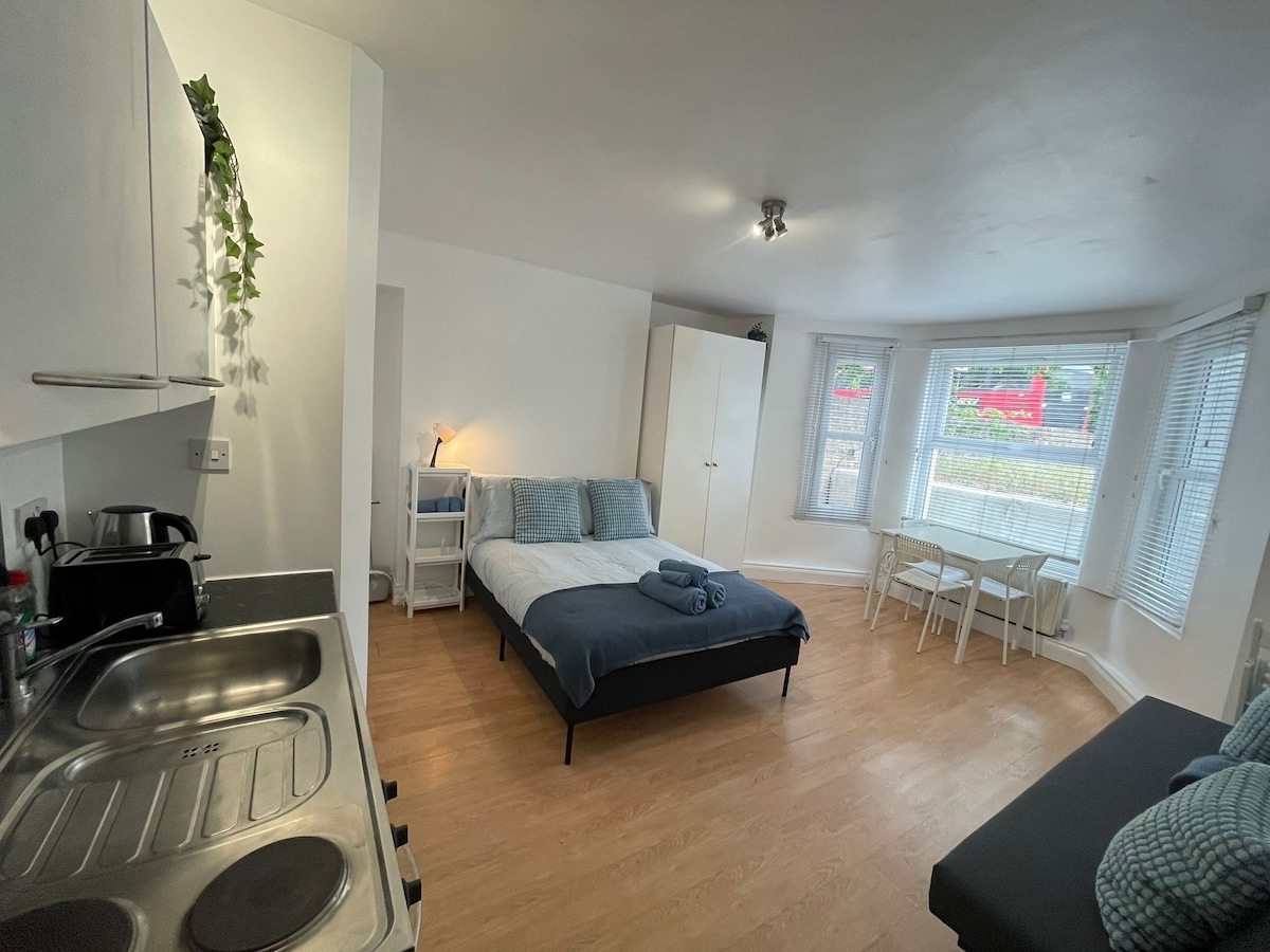 A spacious studio layout features a comfortable sleeping area with a large bed, complemented by soft linens. A kitchenette is visible, along with a dining table for two and cozy seating. Natural light floods the space through bay windows adorned with white blinds.