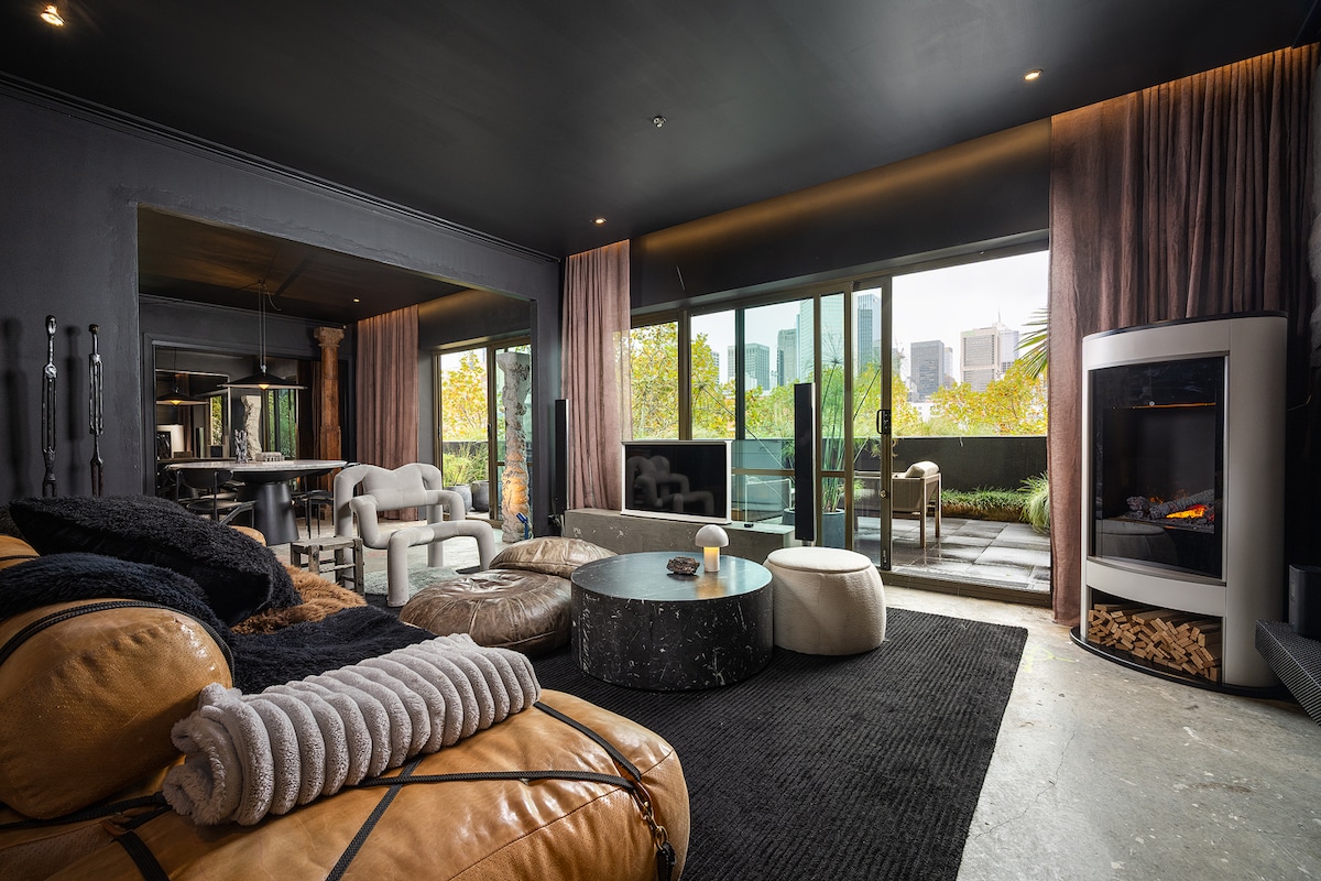 A stylish living area features a tan lounge with various textures, complemented by a sleek black coffee table. Floor-to-ceiling windows reveal city views, and warm natural light enhances the space. A cozy fireplace adds to the ambiance, surrounded by modern furnishings.