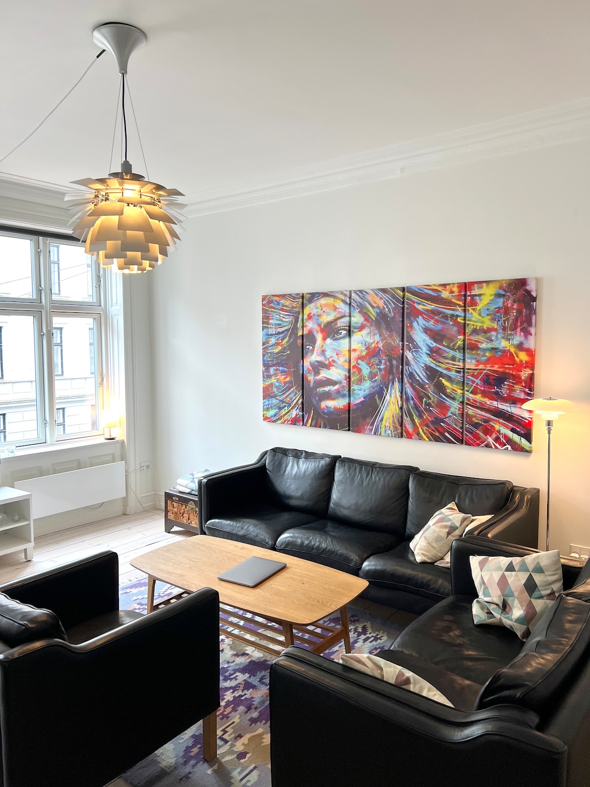 A spacious living area features a black leather sofa set arranged around a light wooden coffee table. A colorful abstract painting is displayed on the wall, complemented by a stylish pendant light overhead. Natural light streams through the large windows, creating a bright atmosphere.