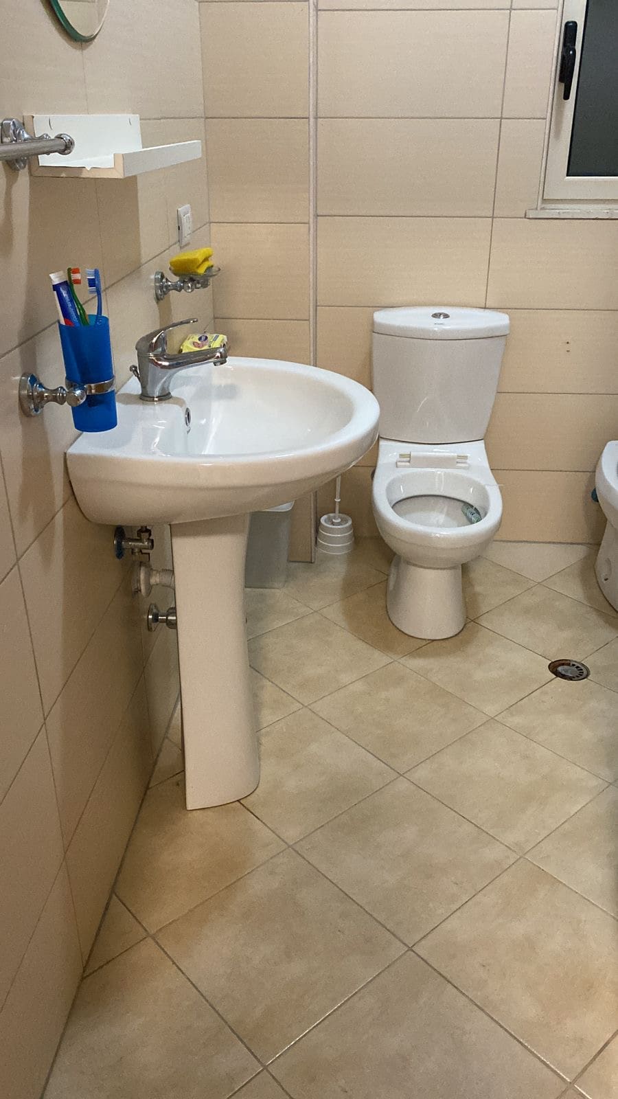 The bathroom features a modern sink with a chrome faucet, accompanied by a discreet shower area. A toilet is positioned beside the sink. Neutral-toned tiles cover the floor and walls, contributing to a clean and functional space. Natural light enters through a small window.