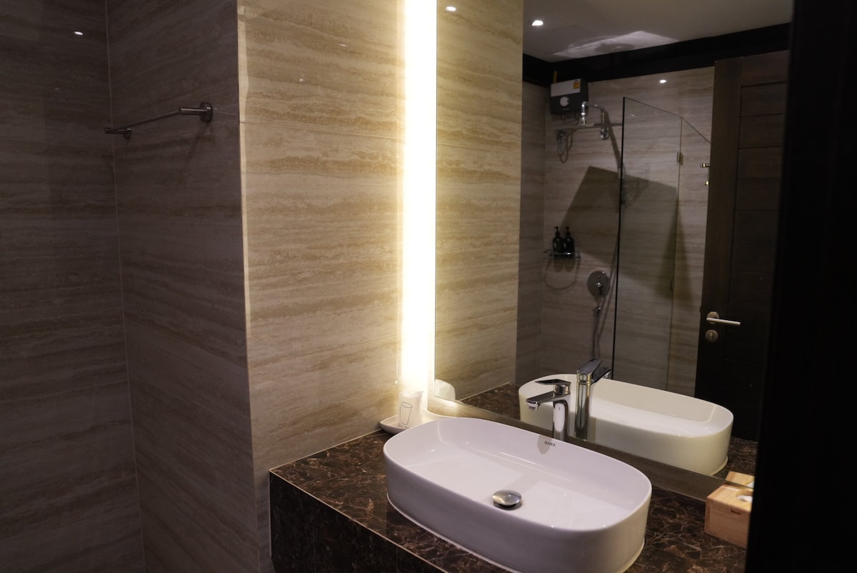 A modern bathroom features elegant stone walls and a marble countertop. A sleek, rectangular sink is positioned under a vertical light, providing illumination. The mirror reflects the spaciousness, while a large shower area is visible in the background, complemented by minimalistic fixtures.
