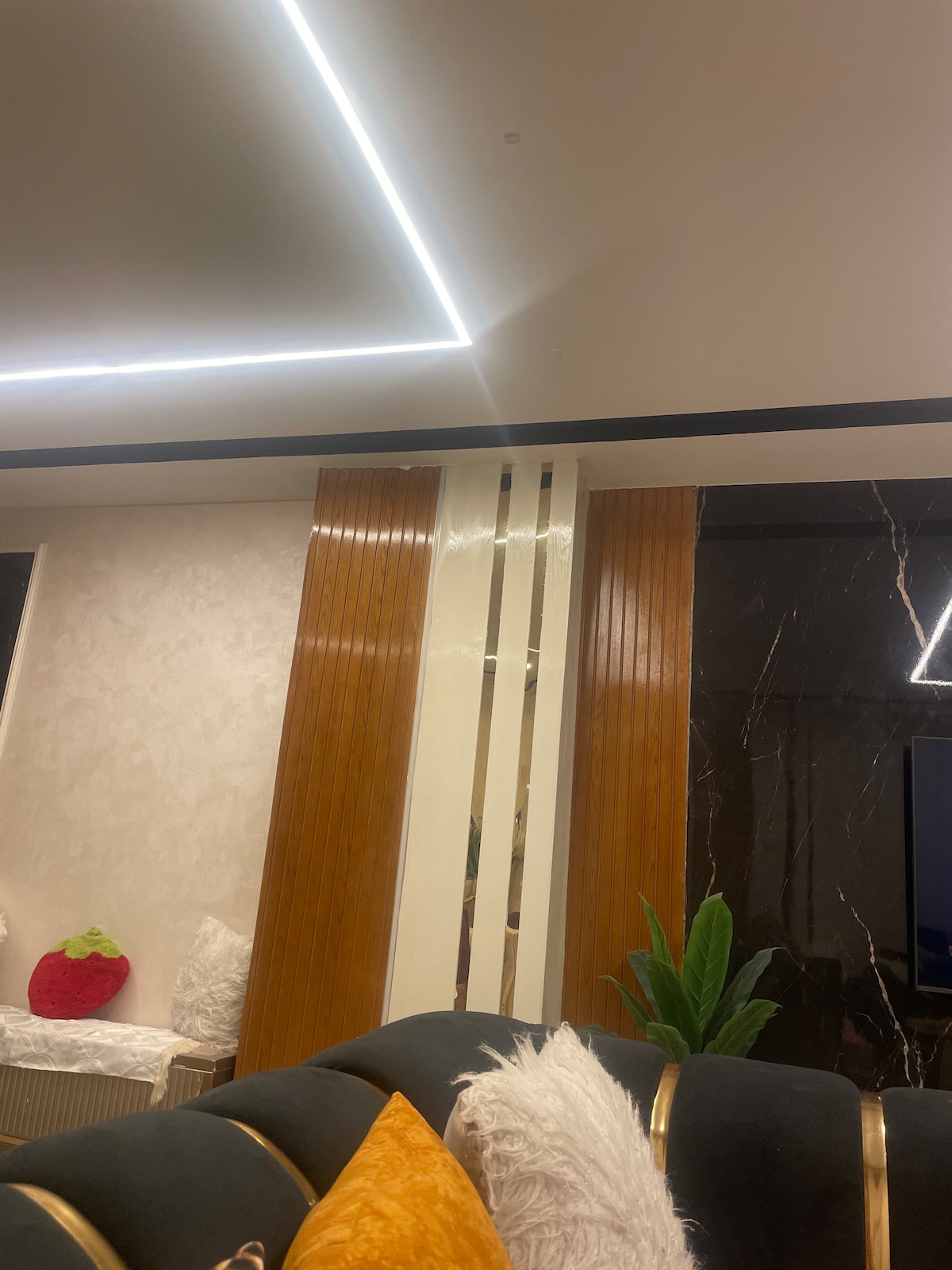 The living area features a dark upholstered sofa adorned with colorful cushions. Two elegant wooden pillars rise alongside a sleek, glossy wall, reflecting soft lighting from modern ceiling fixtures. A green plant adds a touch of nature, complementing the stylish decor.