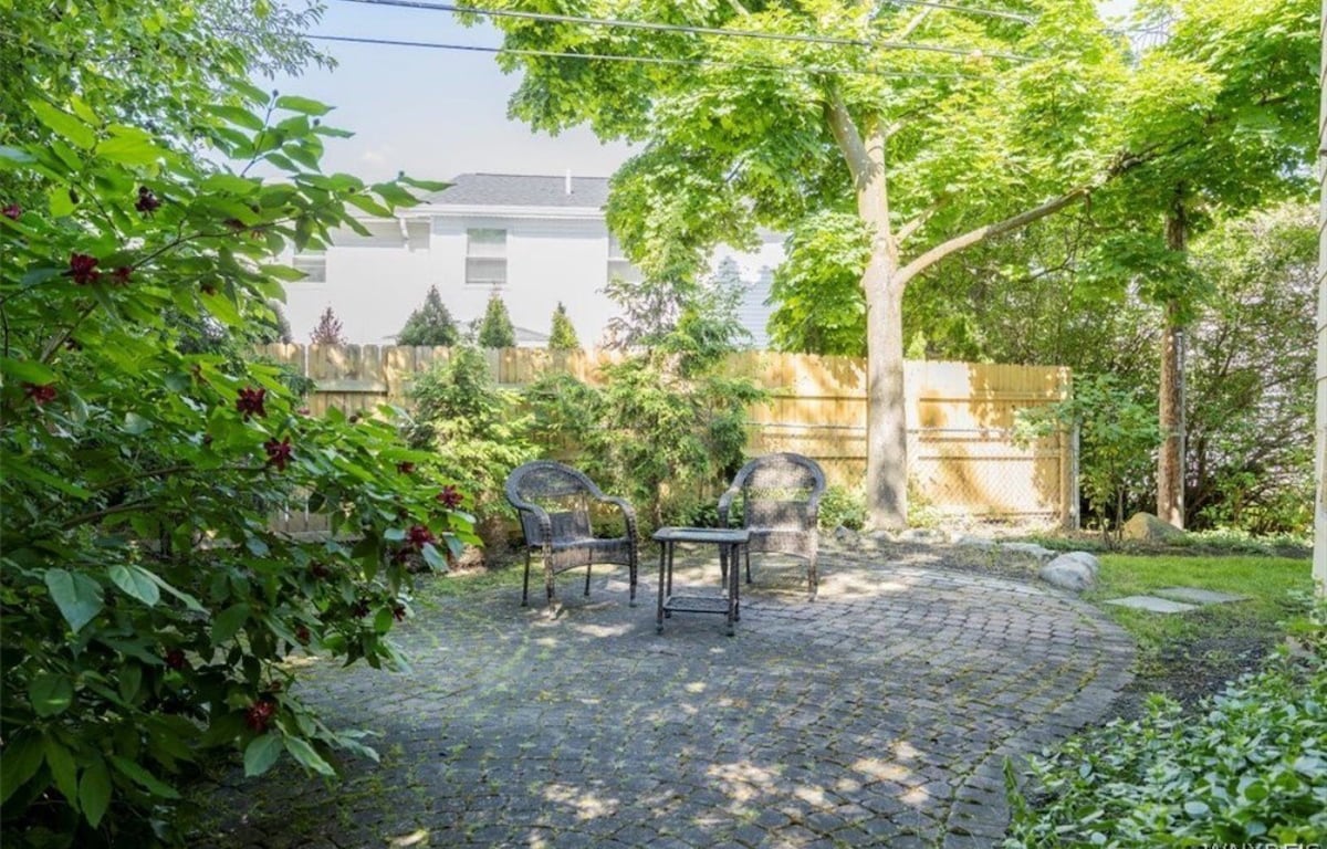 A tranquil backyard space is set with two black wicker chairs and a small table, surrounded by lush greenery. A wooden fence provides privacy, while dappled sunlight filters through the trees, creating a peaceful atmosphere for relaxation.